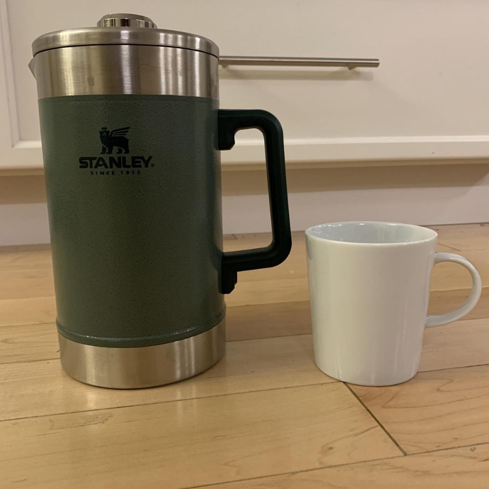 classic green stanley french press sitting on a wood counter next to a white coffee mug.