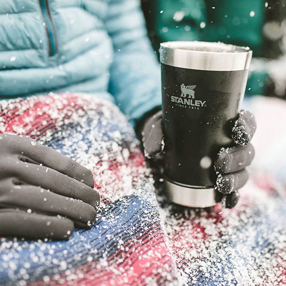 Person sitting  with coat, gloves, and a blanket over their lap, holding a black Stanley beer pint.