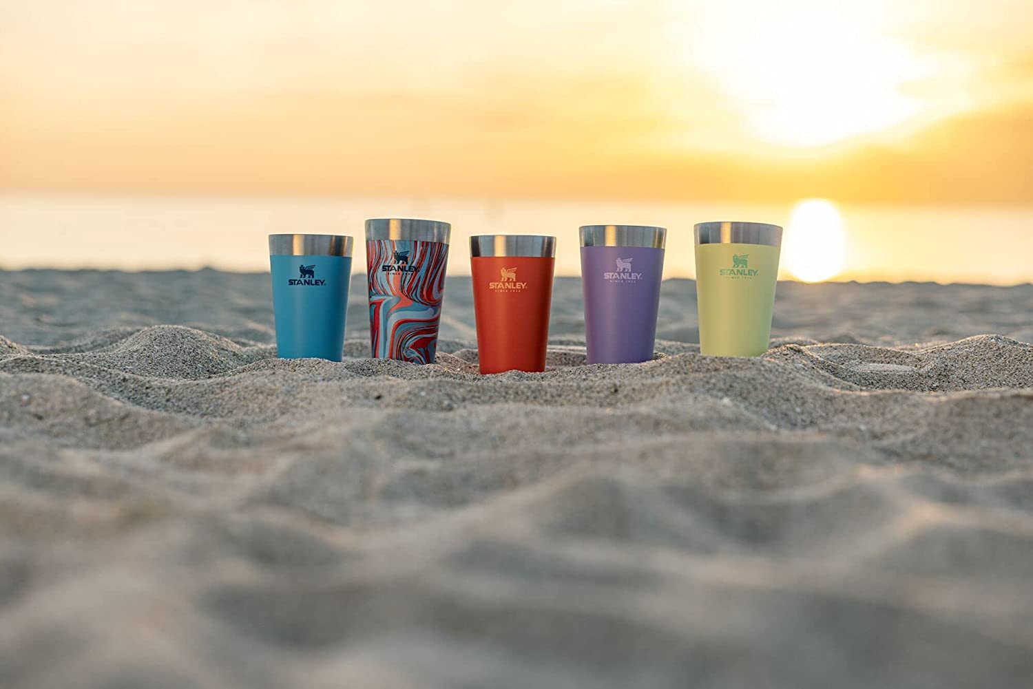 5 Stanley beer pints sitting on the beach with a sunset behind them.